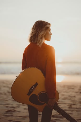 Die Fotografie zeigt eine Frau an einem Strand, die eine Gitarre in der Hand trägt.