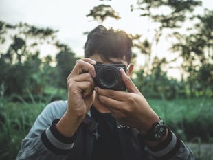 Die Fotografie zeigt eine Person, die eine Kamera hält und diese in Richtung des Betrachters richtet.