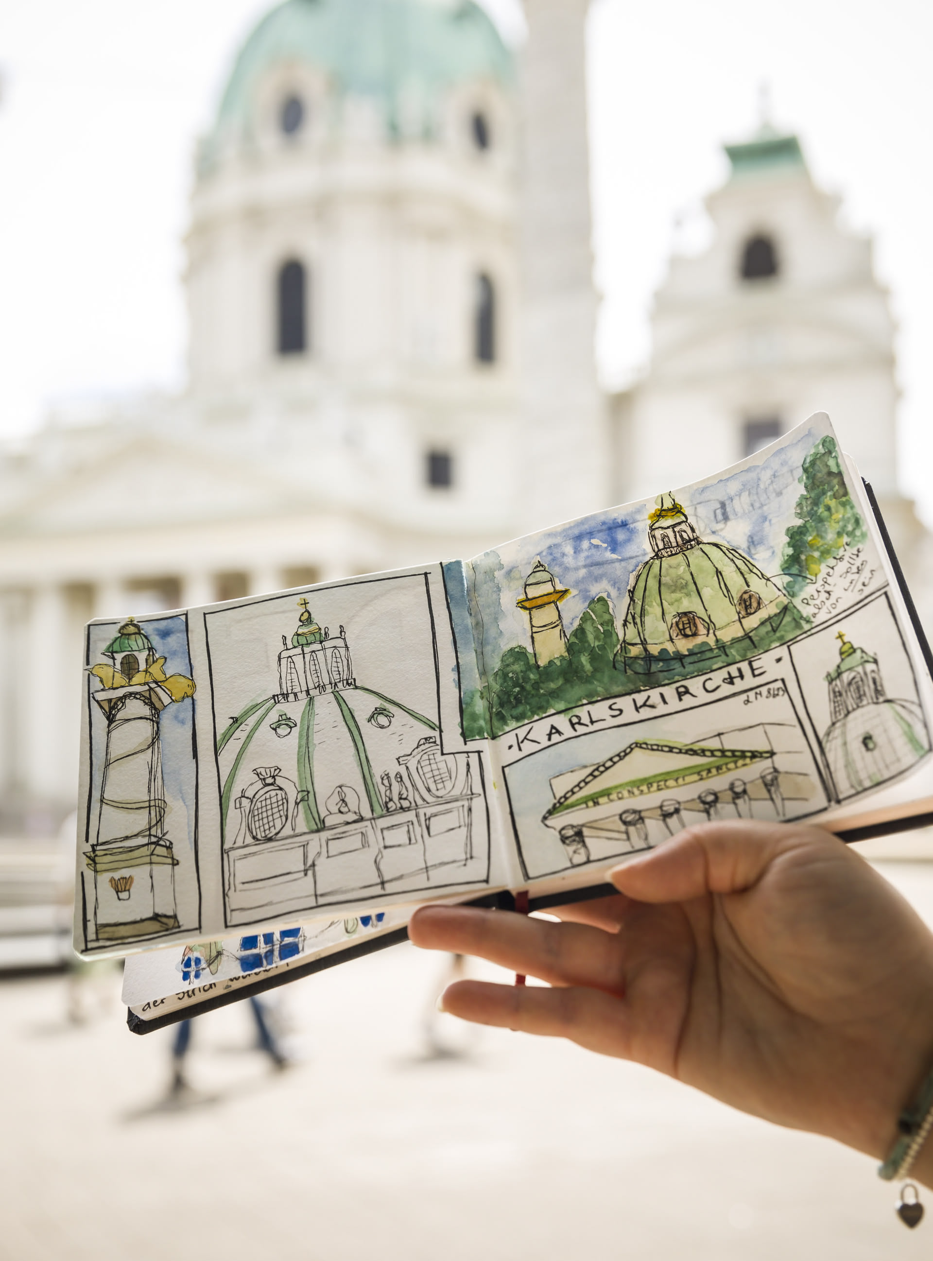 Ein offenes Skizzenbuch mit dem Sketch der Wiener Karlskirche wird draußen vor das Motiv gehalten
