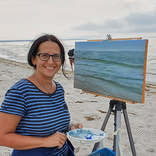 Die Dozentin Elena Romanzin grinst in die Kamera. Neben ihr steht eine Leinwand, auf dem das im Hintergrund zu sehende Meer gemalt ist.