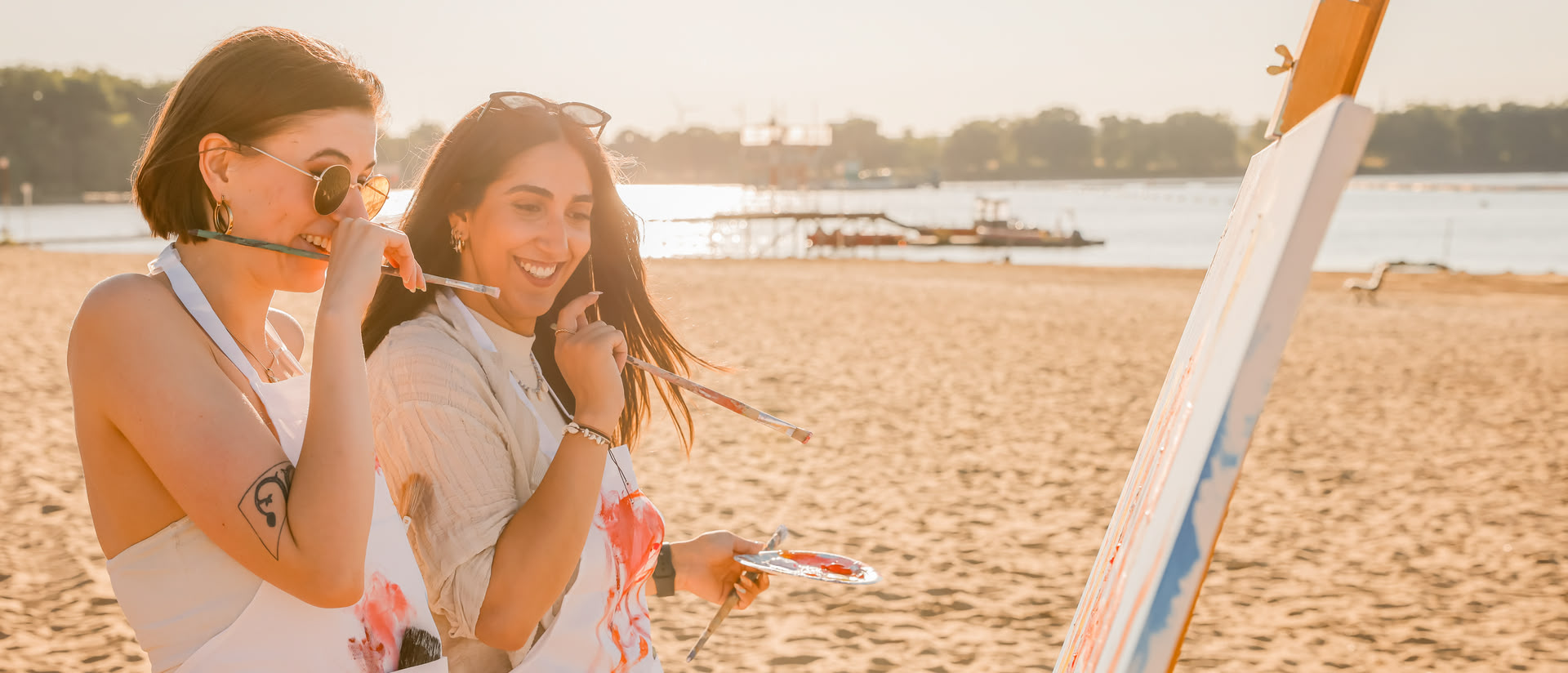 Jara und Tessa von artistravel lachen und blicken auf eine bemalte Leinwand auf einer Staffelei am Strand