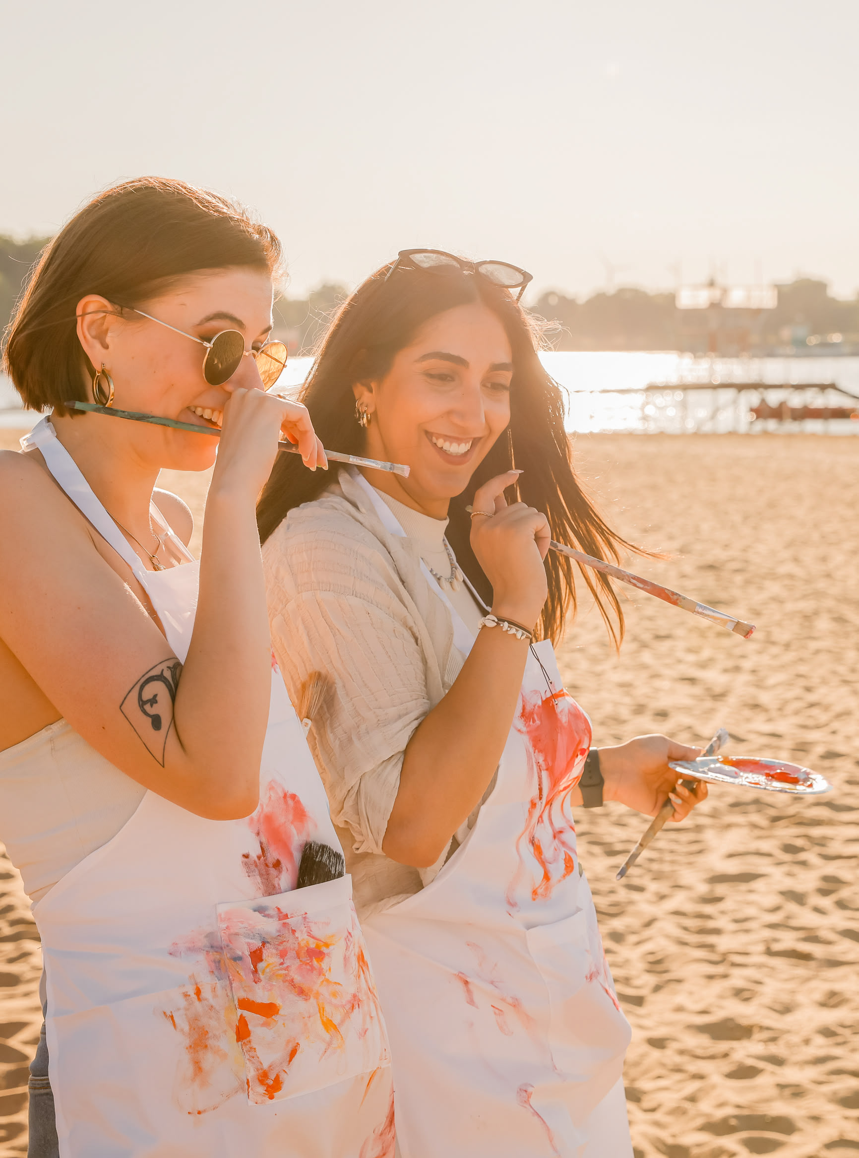 Jara und Tessa von artistravel lachen und blicken auf eine bemalte Leinwand auf einer Staffelei am Strand