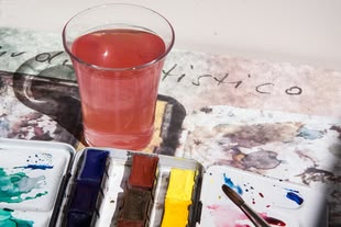 In der Mitte steht ein Glas Wasser, das sich durch die Verwendung von Farbe rot färbt. Im unteren Bereich befindet sich ein Aquarellkasten. 