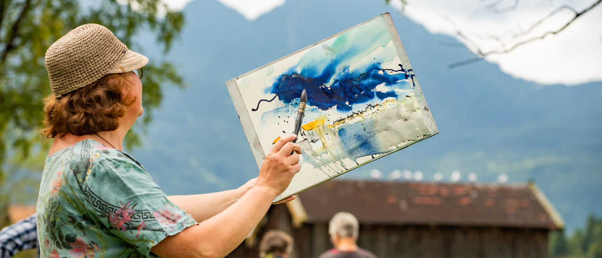 Eine Frau hält einen Malblock mit einem abstrakten Bild darauf hoch und hält einen Pinsel davor, im Hintergrund ist eine Berglandschaft und die Frau blickt in diese Richtung