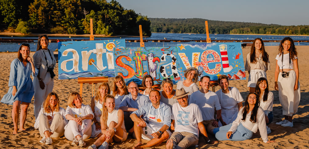 Gruppenbild des artistravel-Teams am Strand vor 3 Leinwänden, auf die das artistravel-Logo gemalt wurde