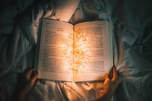 Schreiben lernen - Creative Writing/ Kreatives Schreiben. Foto: Nong Vang / Unsplash Blick von oben auf ein aufgeschlagenes Buch mit einer Lichterkette darin, das von 2 Händen festgehalten wird und auf einem Bett liegt