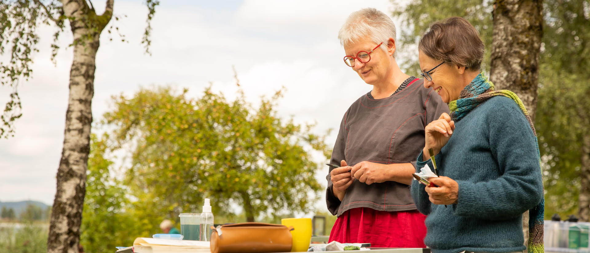 Eine Dozentin (Jutta Höfs) und eine Kursteilnehmerin stehen draußen vor einem Tisch, auf dem Malutensilien liegen