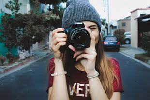 Die Fotografie zeigt eine Person, die auf einer Straße steht und eine Kamera hält und diese in Richtung des Betrachters richtet.