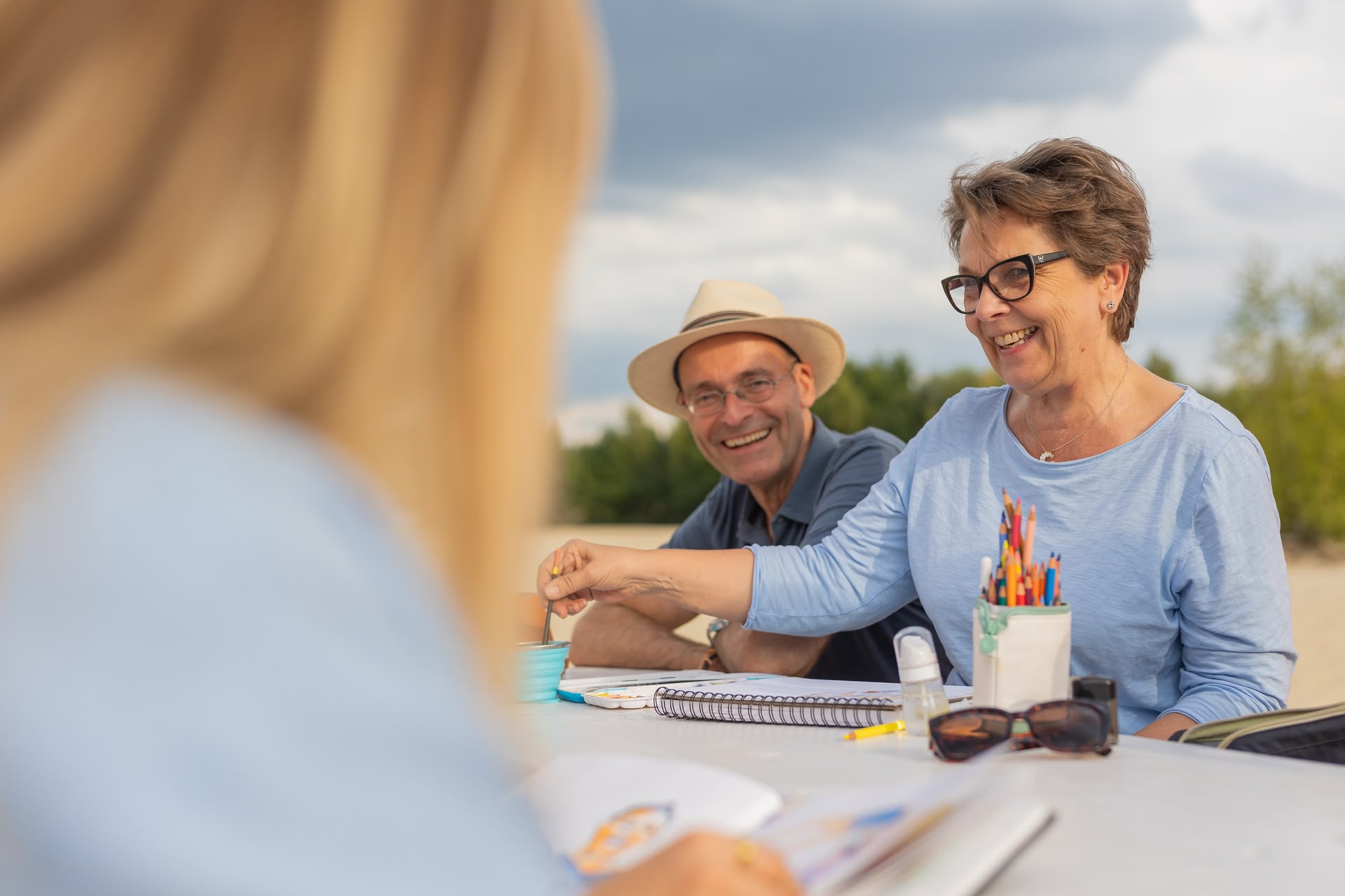 Ein Mann mit Hut und eine Frau (Sebastian und Ulrike) sitzen an einem Tisch am Strand und lächeln