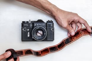 Auf einer weißen Oberfläche liegt eine Kamera, darunter ist eine Hand zu sehen, die einen Filmstreifen hält.