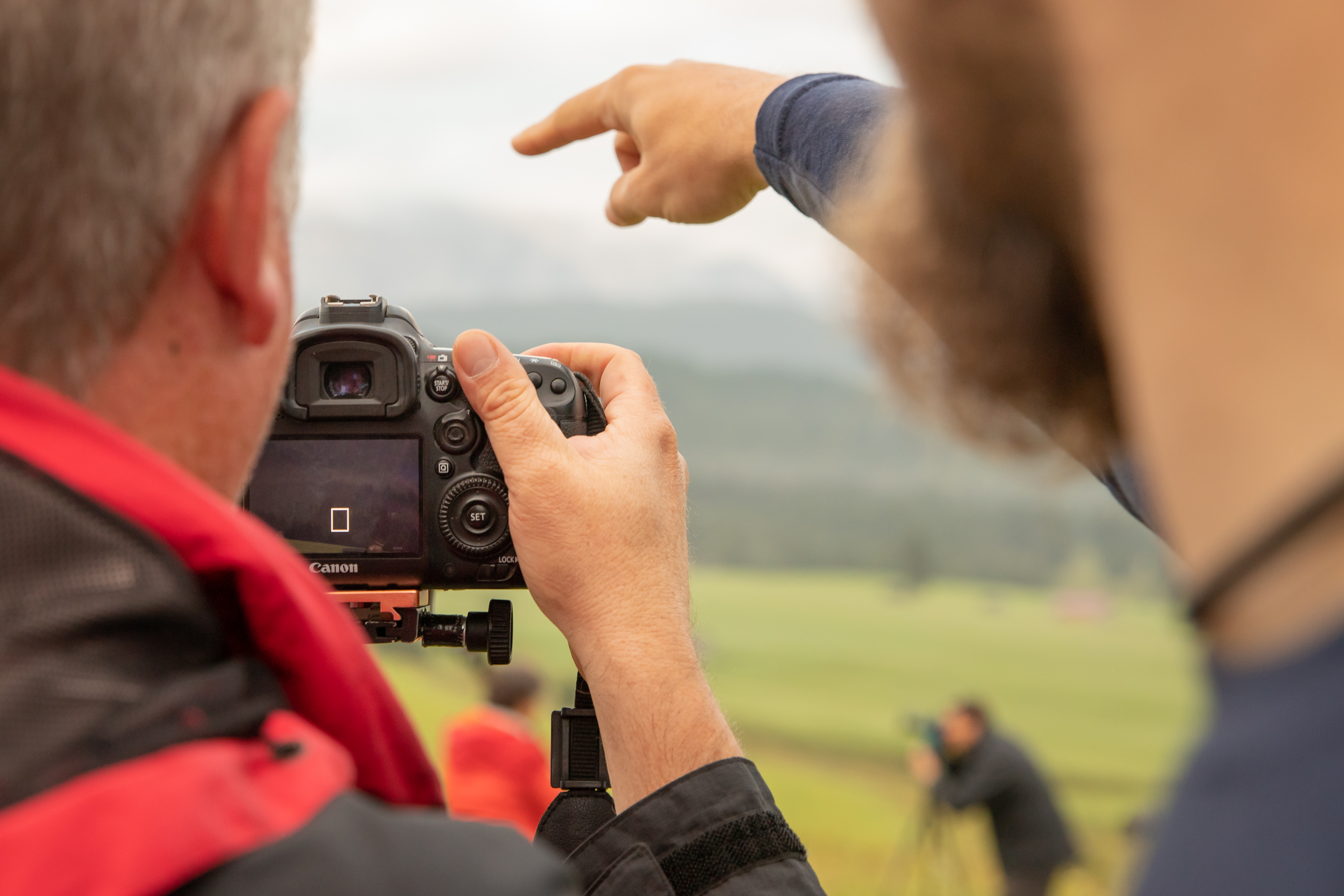 Blick von hinten auf 2 Männer, einer ist Teilnehmer in einem Fotokurs und hält eine Kamera, der andere ist Dozent und erklärt ihm etwas