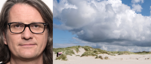 Ein Mann mit langen Haaren und Brille (Foto-Dozent Ralf Niemzig) neben einem seiner Werke (Foto von einer Dünenlandschaft)