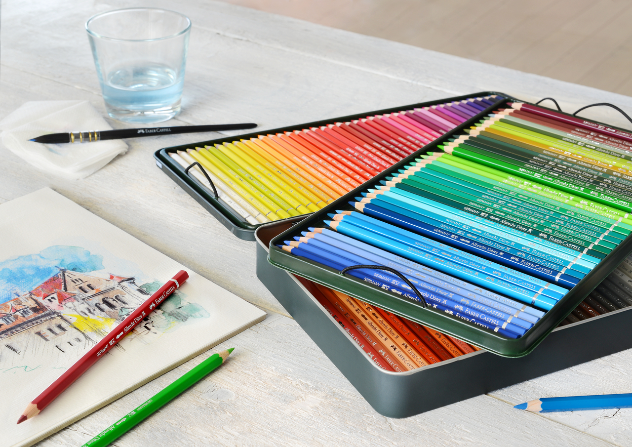 Künstleraquarellstifte von Faber-Castell nach Farben sortiert in einer Metallbox auf einem weißen Tisch, auf dem noch ein Pinsel, ein Glas Wasser, einzelne Stifte und ein gemaltes Bild liegen
