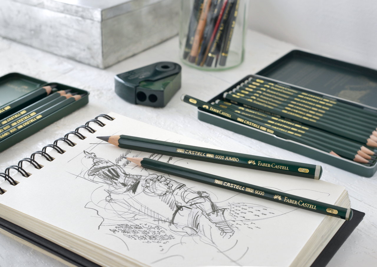 Ein Zeichenblock mit 2 Bleistiften von Faber-Castell liegt auf einem Tisch, daneben zwei Metall-Etuis mit Bleistiften und ein Anspitzer.
