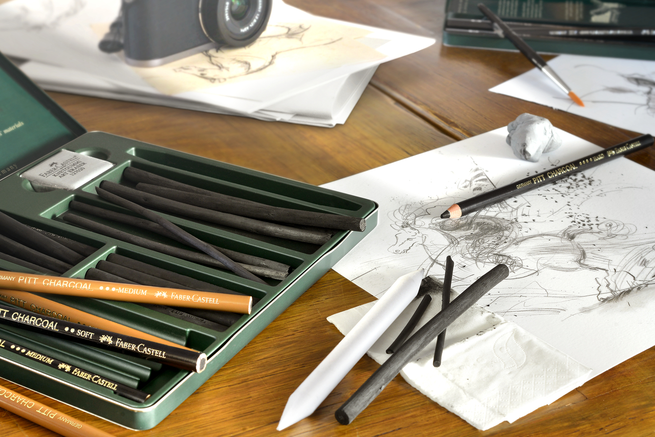 Ein Pitt Charcoal Set von Faber-Castell liegt auf einem Holztisch, daneben eine Kohlezeichnung, die mit diesen Stiften entstanden ist sowie eine Spiegelreflexkamera.