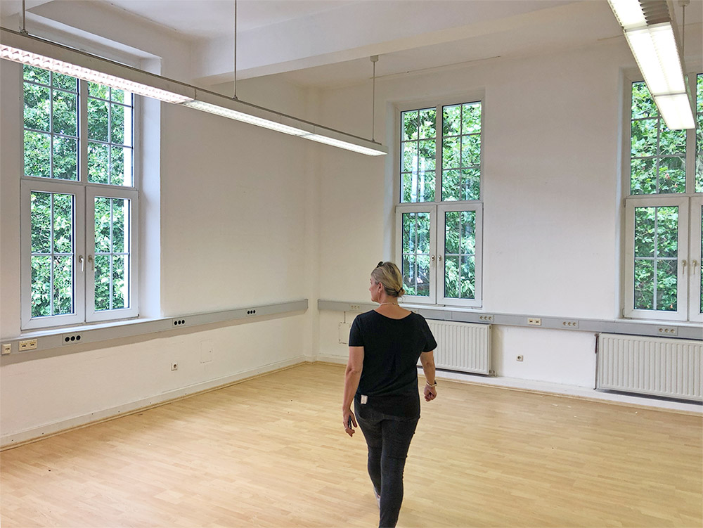 Eine Frau mit schwarzer Kleidung läuft mittig in einen leeren Raum mit Holzboden, weißen Wänden und 3 Fenstern, Ansicht von hinten