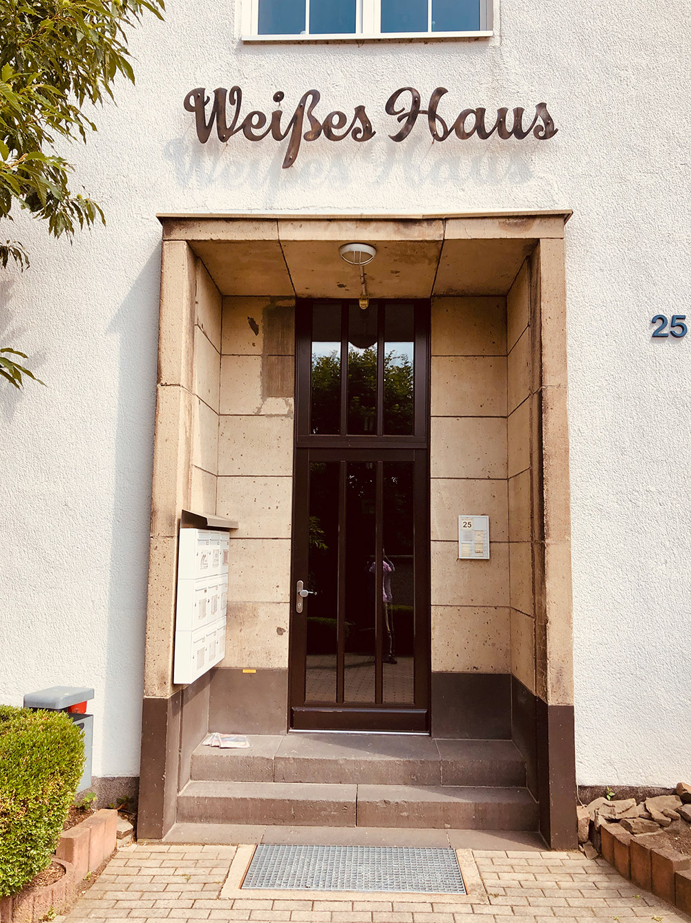 Eingang eines Hauses mit weißen Außenwänden, über der Tür steht "Weißes Haus" in Schreibschrift