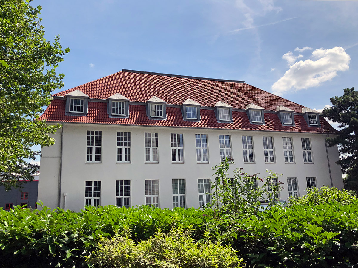 Ein großes weißes altes und gepflegtes Haus mit vielen Fenstern, rundherum sind grüne Bäume und der Himmel ist blau
